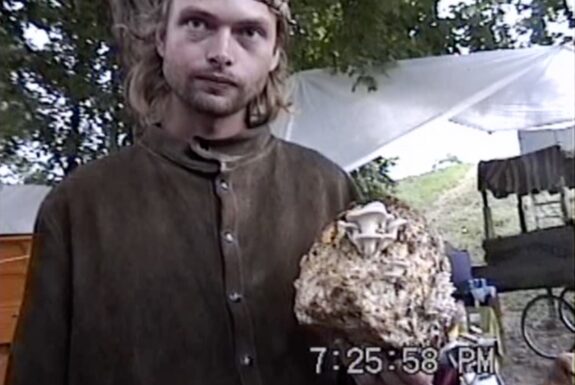 A young man with long blond hair and a beard in a buttoned up collarless brown shirt holds a round object in his hand with mushrooms sprouting out of it.