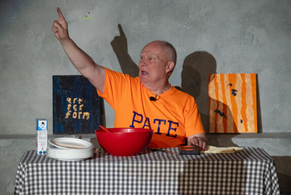 Andrew Paterson, an older man with receding hairline and white eyebrows, sits behind a table that has been covered with a gingham tablecloth. he is looking to the side and speaking, one arm raised with the index finger pointing up. He wears an orange t-shirt with the word PATE in blue letters on the front. Placed on the table are a box of salt, a stack of paper plates, a water glass, a red bowl with the handle of a red implement visible, a black videocassette, and a folded piece of cloth. he is flanked by two paintings. One painting has the words "art per form" in gold against a blue and black background. The other has several black blobs, like large paint drips, against a background of rough vertical strips of orange and gold.