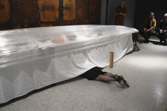 A pair of women's legs, face down, protrude from a long table set with white linens. The legs are dressed in black stockings, black shoes, and the lower part of a knee-length black skirt or dress is also visible. Whatever sits on top of the table is covered in layers of transparent plastic wrap. There appear to be napkins and some a number of brightly coloured objects, perhaps cakes. Some audience members are sitting and standing to one side looking at the legs.