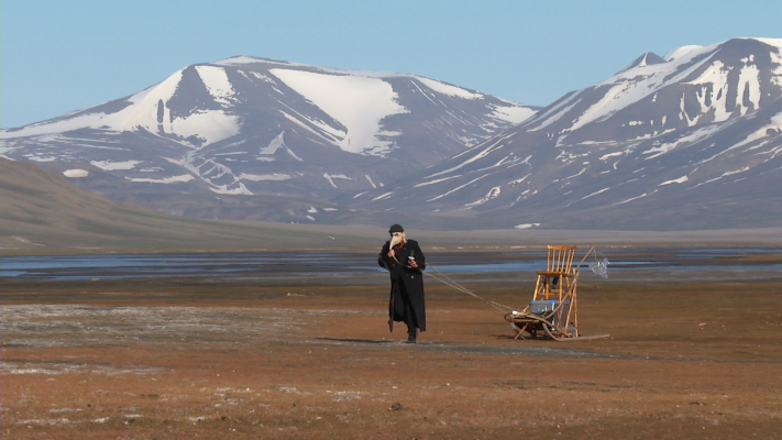 A figure in a dark cloak with a beak mask stands in a barren landscape. She is pulling a sled behind her with long ropes. The sled has a wooden chair and a blue plastic box on it. In the background, mountains with partially covered snow rise in the distance.