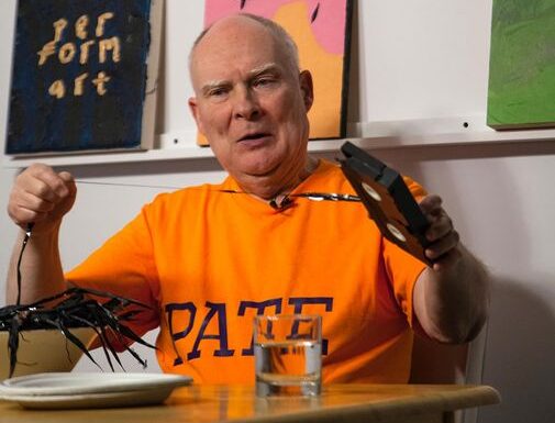 The artist, dressed in an orange shirt with the word PATE on the front, sits at a table set with a small stack of paper plates and a glass of water. He is pulling the magnetic tape out of a VHS video casette. Behind him on a shelf are three unframed abstract canvases.