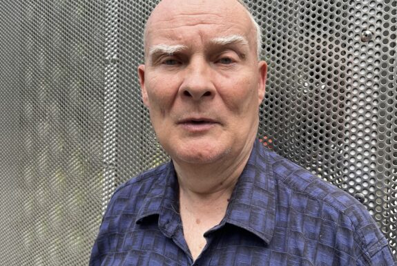 A man stands in front of a metal grillwork facing the camera, his head and upper torso visible. He wears a short sleeve casual shirt with a blue and black pattern of squares. He is clean shave, mostly bald with a fringe of white hair above his ears, and bushy grey eyebrows.