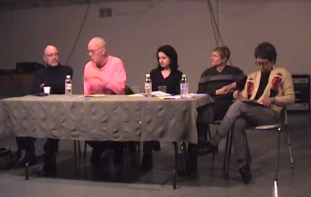 Panelists Bruce Barber, Clive Robertson, Alissa Firth-Eagland, Tangy Duff and moderator Johanna Householder sit at a long table conversing as part of the panel Images versus Iconoclasms or shooting sacred cows from a train of thought, 2002
