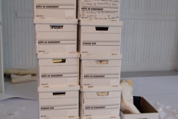 Thre columns of banker's boxes stand side-by side. The largest column on the left is seven boxes tall. The middle column is 6 boxes tall. The column on the right is only two boxes tall. The top box in the left column is open, with paper materials protruding. Various other paper and plastic materials are visible surrounding the columns of boxes. A glass mug sits on the floor in front of the middle column.