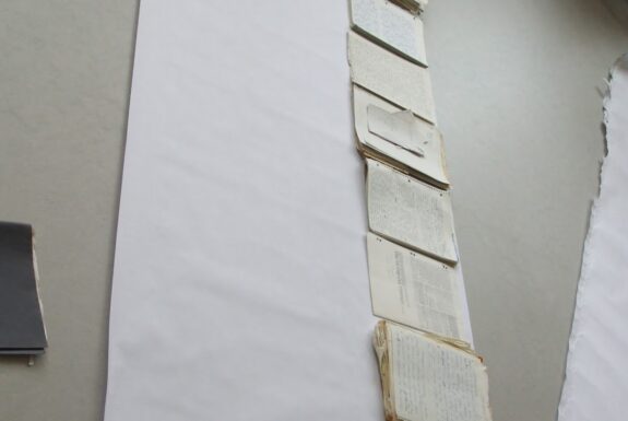 A long roll of white paper is laid out on the floor. Neat stacks of paper, mostly white, some lined, some with hole punches in one side, and some coloured, but each with printing or writing on them, are laid out in a line along one edge of the long white paper roll. The stacks vary in height from a few pages to several inches.