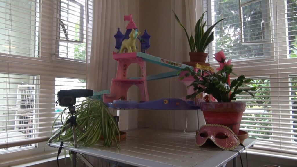 A folding table is set in the corner of a room. Behind the table are windows treated with Venetian blinds and white curtains. Sitting on the table is a colourful My Little Pony toy playground, with a pony on the top tier facing a slide. Houseplants surround the playground. A pink eye mask with gold trim is propped against one pot. A microphone on a table stand also sits on the table pointing toward the playground.