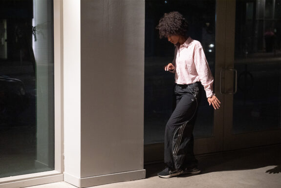 Jessica Karuhanga stands in profile beside a set of glass doors. Her head is tilted downward, eyes almost closed, one arm bent in front of her and the other reaching down and slightly behind, Her fingers are carefully articulated. Her back leg is turned inward, her front leg facing forward to form a perpendicular angle. She wears runners, loose dark track pants with ties at the ankles, and a light top with vertical red stripes. Hes skin tone is tan, her dark hair in an afro.