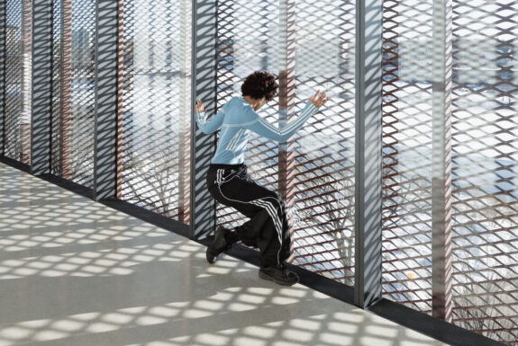 The artist is dressed in athletic wear, pale blue top and black pants, both with white stripe accents, and black running shoes. They stand partially crouched, their back to the camera, head lowered. They use their hands and arms for partial body support, leaning against a glass wall that opens to the exterior. On the other side of the glass, the wall is covered with a metal grill work. Beyond the grill is an urban winter landscape.