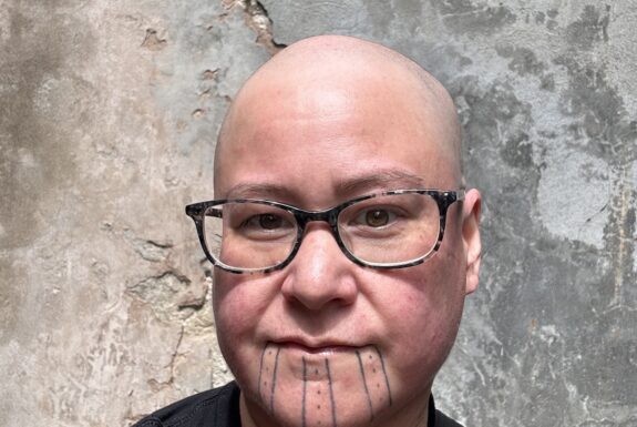 Head an shoulders of a man standing in front of a concrete wall. The man is bald, wears glasees, and has a tattoo with geometric patterns of lines and dots on his chin. Part of another tattoo is visible on their chest above the collar of their black t-shirt.