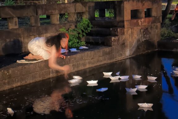 A barefoot figure in a white dress kneels in front of a pool of water at night. A blurry reflection of their form is visible on the surface of the water. They are placing white folded paper boats into the water. A collection of them is already floating.