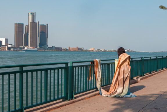 A woman is draped in fabric, her back to the camera. She faces the Detroit River, looking toward a cluster of tall buildings on the opposite shore. The fabric is so large that it hangs down to the ground and is draped over the railing that separates her from the river.