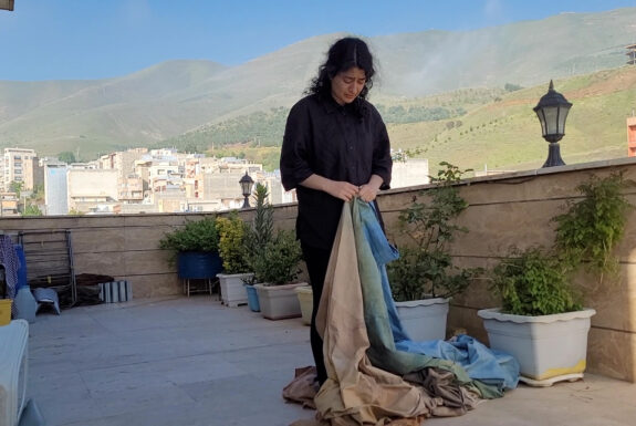 A woman dressed in black stands on a balcony with stone walls. Her head is down. In her hands she holds the edge of a large piece of fabric dyed in pastel browns, greens and blues. Behind her we see apartment buildings and mountains.