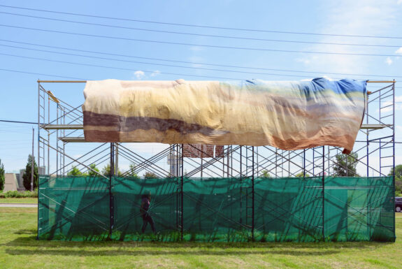 A large piece of fabric in pastel tones of brown, blue, yellow, grey and green hangs from a scaffolding erected on a grassy field. The fabric billows in the wind. A human figure can be seen walking at the bottom of the scaffolding, several feet below the fabric.