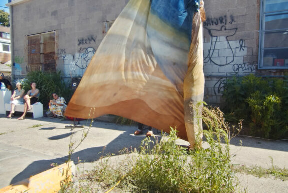 A large piece of cloth, dyed in pastel tones of brown, yellow, grey and blue, billows in the breeze against the backdrop of a cinderblock wall. Part of the fabric is wrapped around something, possible a person. Several onlookers are clustered to the left, some sitting on a white bench and others standing. They stare toward the fabric.