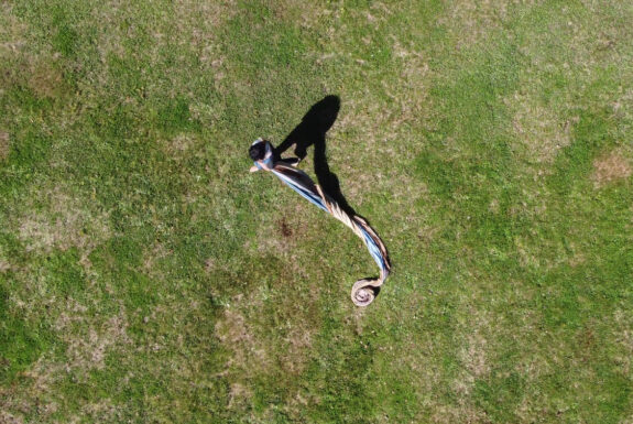 A human figure stands draped in cloth. The fabric trails behind the figure on a grassy ground, curling into a tight spiral. The image is an overhead shot from a drone.