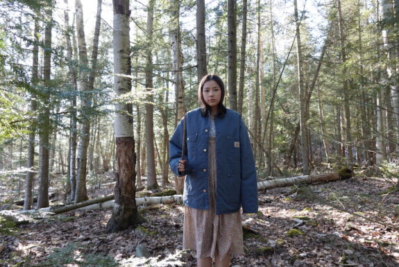 A young native woman dressed in a knee-length coloured dress and a blue jacket stands against a forest backdrop. She holds a stick upright in her right hand as she faces the camera.