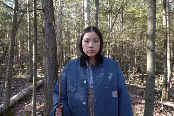 A young native woman dressed in a coloured dress and a blue jacket stands against a forest backdrop. She holds a stick upright in her right hand as she face the camera. It is a close-up image; we see her from the waist up.