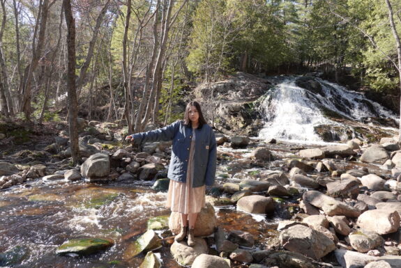 A young native woman dressed in a knee-length coloured dress, a blue jacket, and leather boots stands on rocky ground at the edge of a forest stream, facing the camera. She extends her right arm to the side, over the stream. In her right hand she holds a rock.