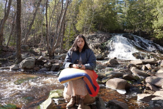 A young native woman dressed in a knee-length coloured dress, a blue jacket, and leather boots sits on a large rock at the edge of a forest stream, facing the camera. In her lap is a coloured blanket. She rests her arms on the blanket, a fist-sized stone nestled in her hands.