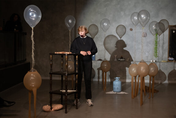 Anja Ibsch a woman with short blonde hair, stands dressed in a black track suit and runners, a thin chain around her neck. in her mouth she holds a large beet. The effort stretches her jaw. Her eyes are closed. In front of her are a set of stacked coffee tables with objects on them. She is also surrounded by several "creatures" each made from two clear plastic balloons filled with helium. The upper balloons of the creatures rise above Anja, forming a head. This is tethered by a furry string to a lower balloon dressed in pantyhose that forms the creatures' torsos. The legs of the pantyhose hang loose blow, just touching the floor.