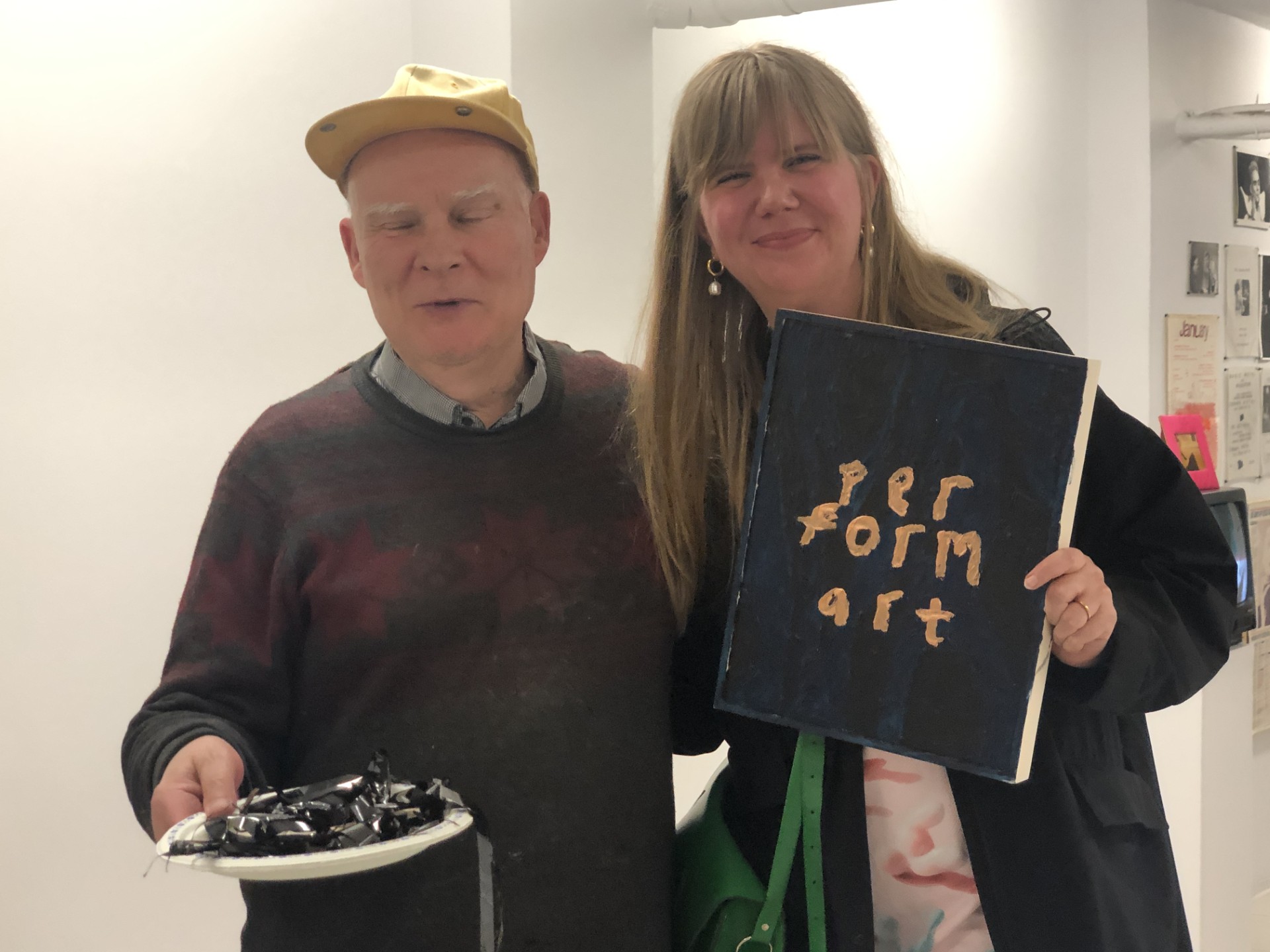 A man and a woman stand arm-in-arm. The man, dressed in a dark sweater and yellow baseball cap, holds forward a paper plate piled with loose videotape. his eyes are closed. The woman holds a painting with the words per / form / art. She is smiling.