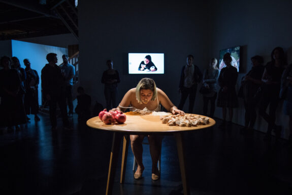 The artist, with blonde, shoulder-length hair, sits at a lit round table, leaning over small mounds of chili powder and curry. Some of the spice coats their lips. On the foreground of the table are piles of pomegranates and eggshells. Above their head is a video screen or projection. In the background, silhouetted audience members watch the artist.