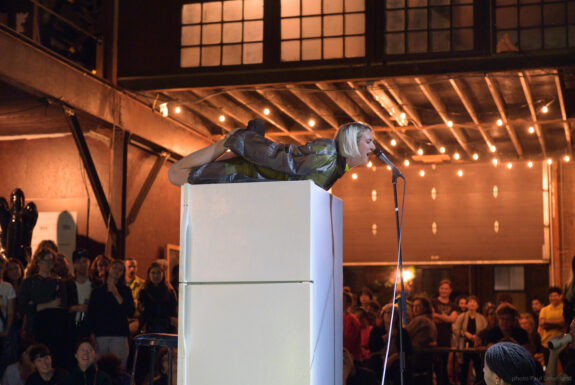 A blond woman lies on her stomach on top of a large white refrigerator. in what appears to be an industrial space with steel beams and an unfinished wood ceiling. She is wearing a transparent plastic raincoat over a neon-coloured dress. Her head is raised and she is reaching her arms back to hold her legs, which are bent at the knees. She is speaking or singing into a microphone on a very tall stand positioned in front of her face. A number of audience members look on from the background.