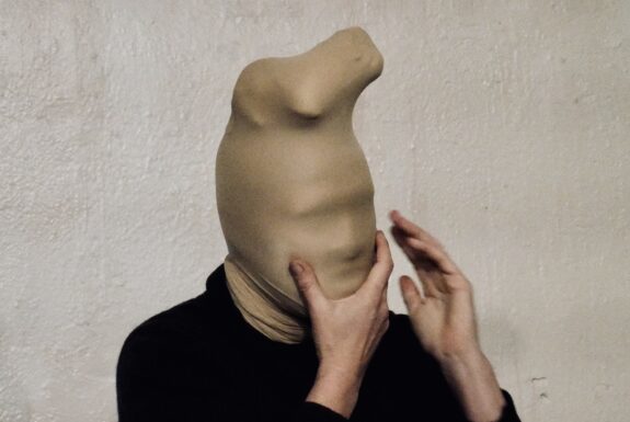 The head and shoulders of a human figure against the backdrop of a white, textured wall. The person's hands reach up toward their head, with the right hand cupping the chin. The head is covered in countless layers of nylon stockings, making the features indistinguishable.