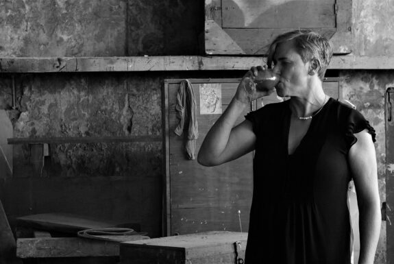 In this black and white image, a woman with short hair in a dark dress drinks a thick dark liquid from a brandy snifter. She is in a room that has an industrial or warehouse feel, with stone walls and wooden boxes covered in dust piled about.