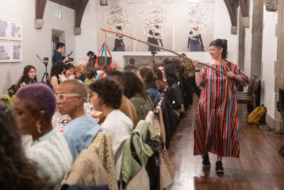 Jess Dobkin, dressed in a shiny dress of colourful vertical stripes and dark runners, walks beside a long table of seated audience members. In her hands she holds a long stick that reaches above the audience members' heads toward the middle of the table. A bucket dangles from the end from colourful macramé strings.