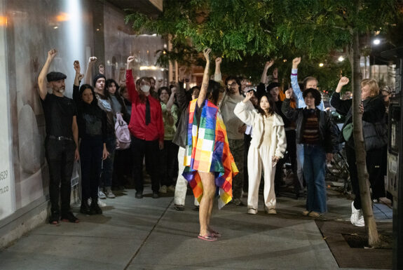 Juma Pariri stands on an evening urban sidewalk, her body draped with a fabric with a pattern of colourful squares that hangs to her knees. Her bare arm and legs are inked with geometric patterns. She wears pink flip-flops. She holds one arm high above her head, a small object in her hand. A crowd of audience members stands in a semi-circle around her. Many also have raised fists or hands with small objects in them.