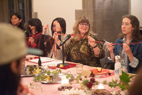 Laurel Green sits among the audience members at an ornately decorated table. Her blonde hair falls below her shoulders, she wears glasses and a glittering gold and brown buttoned Shirt. She is looking to the side and smiling. In her outstretched hands she holds a segment of red yarn. The audience members on either side of her also hold lengths of the yarn.