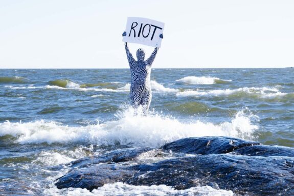 A human figure in a full-body zebra patterned costume stands in the waves of a vast body of water. They hold a placard above their heads that has the word RIOT printed on it.