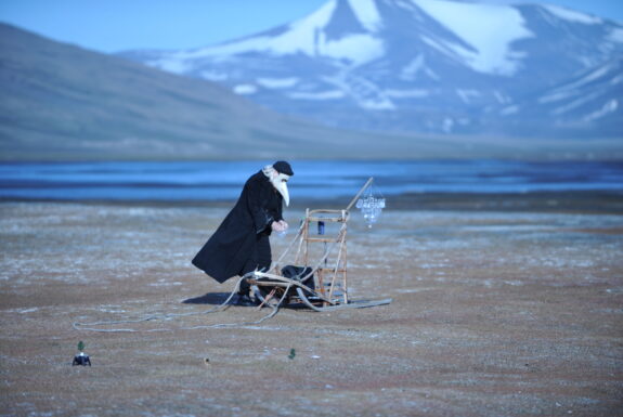 A figure stands leaning over a sled with a raised back frame. The sled has a crystal chandelier hanging off the back. The figure is dressed in a long black coat, black boots, and a black beret. Their hair is grey and their face is covered by a white Commedia dell'arte-style mask with a long beak. The hold something in their hands. Around them is a barren landscape, gravelly earth with a a body of water and mountains covered in patches of snow rising behind the figure in soft focus.