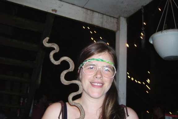 The artist is facing the camera, smiling, visible from the shoulders up. They have long brown hair, glasses, and a thin crocheted headband in shades of green and white. In one hand they hold a curvy wooden stick with a shape evocative of a snake.