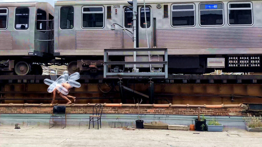 A naked female figure, covered in clear plastic balloons, is moving on all fours along the top of a low brick wall. Behind her are two connected cabins of a silver subway train. On the concrete foreground are some mismatched chairs and a number of planters, some with living plants in them.