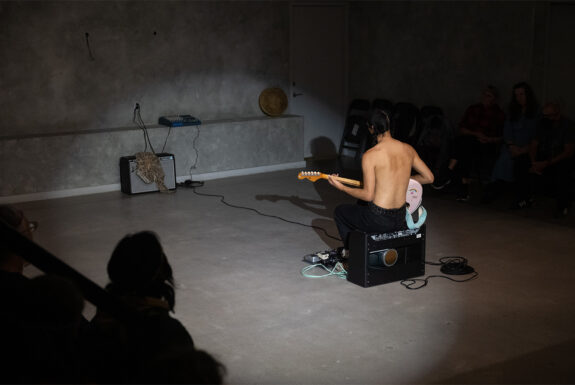 seth cardinal dodginghorse sits on a black amplifier with their back to the audience. They have dark hair with two braids, they are shirtless, and they are playing a pink electric guitar. They sit facing a second amplifier with a shirt draped over the front of it.