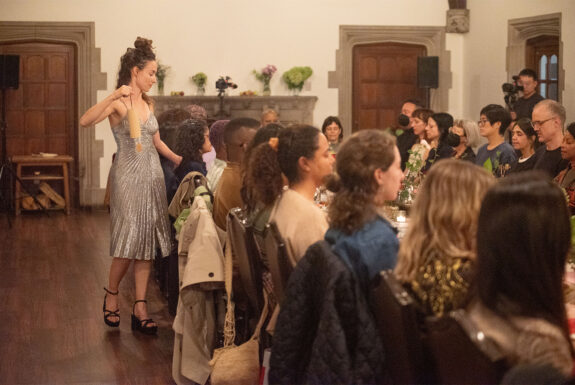 Shalon T. Webber-Heffermam stands beside a long table with audience members seated around it. She is dressed in a tightly fitted sliver pleated evening dress and black wedge-heeled sandals. Her long brown hair is piled high in the front and hangs loose at her back. In one hand, she holds up a cylindrical wooden bell. The other hand is placing something onto the table in front of one of the audience members.