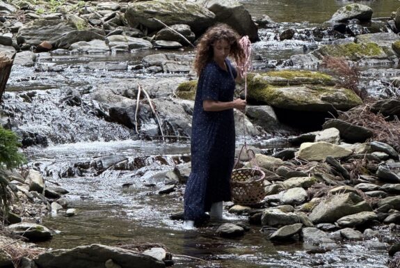 A woman in a long blue summer dress stands in a rocky stream. She is carrying a basket of rocks that hangs from a long rope.