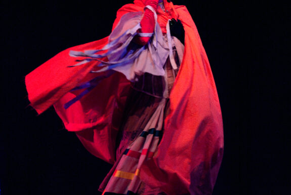 The artist stands against a black backdrop, facing the camera. They are dressed in a flowing dress of lavender and pink, with a full-length red cape that billows out around them. They hold one splayed hand, covered in an arm-length red glove, over the lower part of their face, covering their mouth.