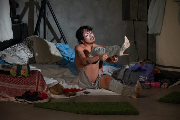 Tyler J. Sloane sits on the floor, surrounded by clothes, fabric, bags and other objects. They have dark hair and a Balbo-style beard. Their face is made up with purple eyeshadow, mascaraed eyelashes, and red lipstick. Thy are shirtless, with short grey shorts, and white stockings that come above the knees. One leg stretches long in front of them, adorned with a silver sequined boot with stiletto heel. They are in the midst of pulling on the second boot, leg held high in front of them. They are shirtless, with large tattoos visible on their upper arm and forearm.