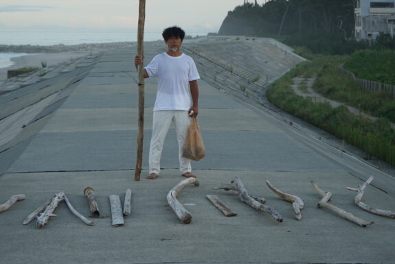 The artist stands on a concrete dam, ocean on one side and young pine tress on the other. He is dressed in a white t-shirt and white pants, barefoot, facing the camera. In one hand he holds a large bamboo driftwood staff, taller than he is. In the other he has a plastic bag, weighted down by whatever is inside it. A number of large pieces of driftwood are placed on the ground in front of him.