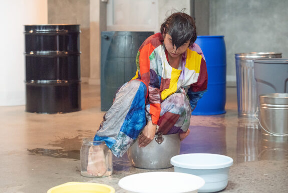 Vanessa Godden sits crouched with one foot in a metal pot and the other in a smaller clear glass jar. Water in the glass jar distorts the look of their foot, making it appear larger. They are barefoot, wearing a matching top and pants with a pattern of colourful abstract rectangles. A thin pool of water surrounds them on the concrete floor. Arrayed in s semi-circle around them are a mismatched series of metal, plastic, and glass containers of gradually decreasing sizes.