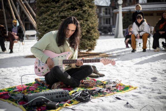 The artist sits cross-legged on a colourful blanket placed on top of snow-covered ground. They wear a light-coloured pullover, dark pants, and moccasins. They are playing a pink electric guitar, with a small mixer and other electronics laid out in front of them on the blanket. In the background are houses, evergreens, and a tipi frame. Several people sit on folding chairs, listening.