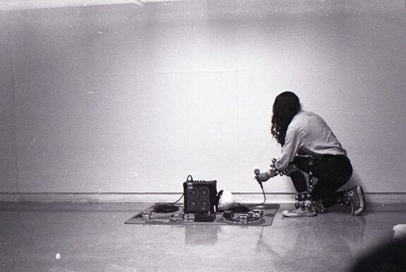 In this black and white image, a long-haired young man dressed in a white long-sleeve shirt, black jeans and running shoes kneels in front of a small blanket set with electronic equipment. He holds a microphone in one hand. His arms and wrists are adorned with strips of dance bells.