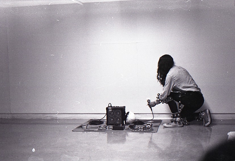 In this black and white image, a long-haired young man dressed in a white long-sleeve shirt, black jeans and running shoes kneels in front of a small blanket set with electronic equipment. He holds a microphone in one hand. His arms and wrists are adorned with strips of dance bells.