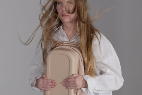 A woman dressed in a white shirt is visible from the waist up. her long strawberry blonde hair is blowing in front of her face. She stares foward with a determined gaze. She is clutching a tan-coloured collapsed plastic bin to her torso.