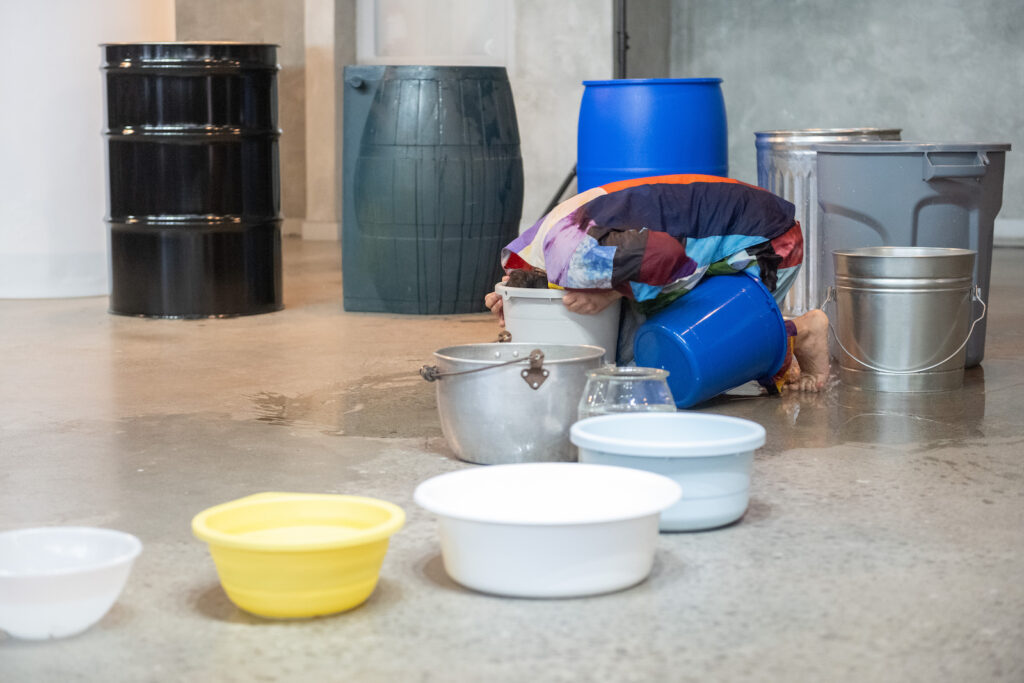 Vanessa Godden is crouched on the floor, feet up on their toes, head immersed in a white bucket. Their hands clutch the rim of the bucket. They are barefoot, wearing a matching top and pants with a pattern of colourful abstract rectangles. A thin pool of water surrounds them on the concrete floor. Arrayed in s semi-circle around them are a mismatched series of metal, plastic, and glass containers of gradually decreasing sizes.