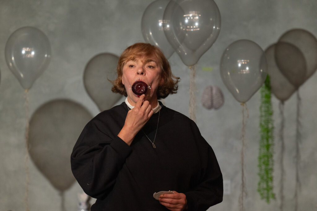 Anja Ibsch, dressed in a black sweatshirt with a white collar underneath and a thin silver necklace with a razor blade on the end, faces toward the camera. A red beet protrudes from her mouth. There are several pins with large round heads stuck into the beet. She is pushing a pin into the beet with the index finger of one hand. her other hand holds a wheel with more pins at waist level. Several clear plastic balloons float around her, with fuzzy strings dangling down, their translucent shadows visible on the wall behind her.