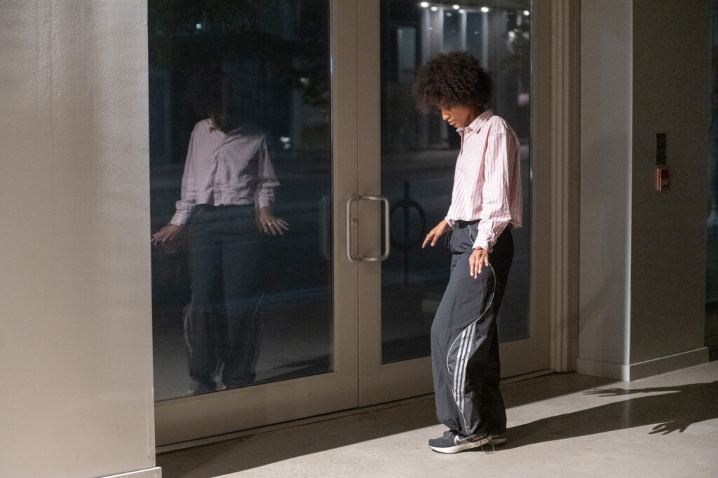 Jessica Karuhanga stands diagonally, facing a set of glass doors. Her head is tilted downward, eyes almost closed, her arms slightly raised above her sides, fingers reaching, one foot barely raised at the heel. She wears runners, loose dark track pants with ties at the ankles, and a light top with vertical red stripes. Hes skin tone is tan, her dark hair in an afro. The side lighting casts a long shadow behind her. One pane of the glass doors reflects a blurred frontal image of the artist. A street-lit urban road and sidewalk are faintly visible beyond.